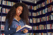 © Seventyfour - Young adult biracial woman writing in notebook while standing in front of bookshelves in library, holding pen and looking at page, long curly hair framing focused expression