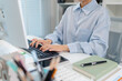 © PicDY - A young Asian businesswoman is sitting at her desk in a modern office, reviewing printed documents while working on a computer, showing focus, dedication, and professional attitude.