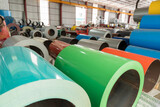 Close-up of large metal coil rolls with brightly colored coatings (painted steel) stacked in a spacious industrial warehouse or factory setting.