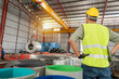 © tong2530 - Industrial worker in safety uniform overseeing the operation of an overhead crane lifting a metal coil roll inside a bright factory warehouse.
