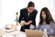 © Studio Marmellata - Two young businesswomen collaborate on a laptop in a modern office. One stands, pointing at the screen, while the other sits, focused on the task. They are engaged in a productive discussion.