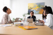 © Studio Marmellata - A diverse group of professional women collaborates in a bright office meeting. They are engaged in discussion, working on laptops, and sharing ideas around a large wooden table.