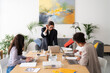 © Studio Marmellata - Three diverse women collaborate in a modern office setting. One stands, adjusting her glasses while reviewing a laptop. Two colleagues work on computers and documents at the table.