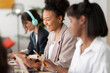 © Studio Marmellata - Diverse young women are working together in a modern office setting. The central woman smiles brightly, holding a pencil and looking at her laptop. Another woman wears headphones, focused on her task.