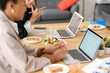 © Studio Marmellata - Two diverse women work on laptops at a shared desk, enjoying a healthy lunch of salads and sandwiches. They are focused on their tasks in a modern office setting.