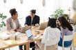 © Studio Marmellata - Four diverse young women are collaborating at a bright office table. They work on laptops, share ideas, and engage in cheerful discussion during a productive meeting.