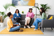 © Studio Marmellata - Four diverse young women are happily chatting and smiling in a modern living room. They are enjoying a friendly gathering, sitting on a sofa, armchair, and floor.