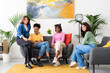 © Studio Marmellata - Four diverse young women sit comfortably in a modern living room, looking at the camera. They are friends enjoying a relaxed gathering at home.