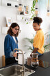 © Studio Marmellata - Two young women prepare coffee in a modern kitchen. One fills a kettle while the other sits on the counter, smiling. They enjoy a casual morning together.