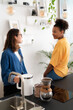 © Studio Marmellata - Two diverse young women share a moment in a bright kitchen. One prepares coffee with a kettle while the other sits on the counter, engaging in friendly conversation.