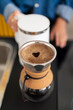 © Studio Marmellata - A person in a blue shirt and yellow apron pours hot water from a white kettle into a pour-over coffee maker, creating a heart shape in the coffee foam.