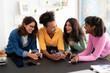 © Studio Marmellata - A group of four cheerful young women are enjoying a casual gathering at home, sharing laughs and looking at a smartphone while drinking beverages.