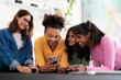 © Studio Marmellata - Four diverse young women gather around a table, smiling and looking at a smartphone. They share a happy moment, enjoying content together while holding drinks.