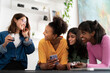 © Studio Marmellata - Four diverse young women are smiling and laughing together, looking at a smartphone and holding drinks. They are enjoying a casual gathering indoors.