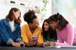 © Studio Marmellata - Four diverse young women friends are happily socializing at home, looking at a smartphone and laughing together. They are enjoying drinks and sharing a moment.