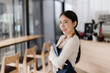 © Tj - Asian woman barista wearing an apron and smiling, holding a digital tablet in a modern coffee shop, portraying small business ownership, hospitality, and customer service