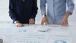 © rogerphoto - Business colleagues are examining financial data, pointing at charts and graphs spread out on a table during a collaborative analysis session in a modern office.