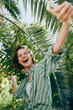 © SHOTPRIME STUDIO - Bright, joyful portrait of a woman outdoors taking a selfie among lush palm leaves, wearing a striped shirt and radiating summer energy.
