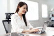 © NAMPIX - A cheerful young Asian businesswoman smiling happily while typing and working efficiently on a laptop computer in a bright, modern office or home setting.