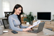 © NAMPIX - A beautiful young Asian woman smiling happily while reviewing paper documents and working on her laptop at a home office desk.