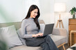 © NAMPIX - A beautiful young Asian woman focused on her work, sitting comfortably on a sofa and using a laptop computer for remote work or online studying at home.