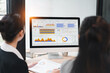 © kenchiro168 - Data visualization dashboard on desktop monitor with two businesswomen reviewing colorful graphs and charts in bright office