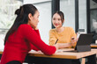 © kenchiro168 - Young woman in yellow sweater showing calculator to colleague while discussing finances at table, friendly collaboration and focused planning