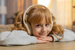 © Andrii Iemelianenko - Young girl lying on table listening to an audiobook with headphones, reacting with excitement, surprise. Caucasian child smiles joyfully, wondering what happens next in story, enjoys adventure at home
