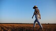 © ExpressVectors - Adult agronomist in straw hat walking among barley plantation at dusk. Female farmer going through wheat meadow at sunset. Beautiful scenic landscape. Concept of agricultural business. Slow motion