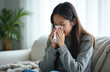 © Pete - Young woman sits on sofa with tissues. She sneezes into paper, feeling unwell with cold or flu symptoms. Home interior provides comfort while she rests.