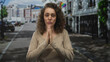 © Krakenimages.com - Young hispanic woman with hands pressed together praying on a city street in front of townhouses and storefronts; pleading hope.