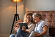 © luciano - Portrait of happy mature blonde curly woman sitting on the floor close to her elderly mother sharing together social news on digital tablet. The scene captures affection, and emotional connection