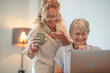 © luciano - Blonde curly woman helping an older woman use a laptop at home while having a coffee break and smiling. The image represents intergenerational connection, technology learning, and family bonding