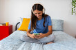 © Xavier Lorenzo - Caucasian teenage girl smiling and looking at her phone while wearing headphones and sitting on her bed. Youth, connection and technology lifestyle concept.