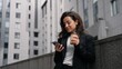 © Vlad - Close up. Caucasian female standing outdoors around grey modern buildings, working on her phone, smiling, looking around, and having drink from cup .