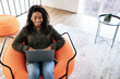 © Prostock-studio - A young woman sits comfortably in a bright orange bean bag chair, focused on her laptop. Sunlight streams through large windows, creating a warm atmosphere in the stylish room.