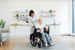 © sofiko14 - A caregiver pushes an elderly woman in a wheelchair, smiling at each other in a bright, modern kitchen setting. The scene conveys care and support.