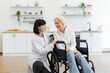 © sofiko14 - A smiling caregiver comforts an elderly woman in a wheelchair, fostering a sense of support and care in a bright, modern home setting.