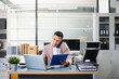 © laddawan - Asian businessman talking on smartphone while working on laptop at modern office desk, symbolizing business communication and corporate success.