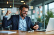 © Vadym - Man in suit celebrates success while looking at smartphone in modern office. He smiles looking at phone with fist raised. Laptop and tablet are on desk. Excitement at work.