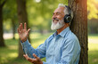 © Vadym - Senior man with white beard listens to music using headphones outdoors. Leans against tree trunk in park, smiling broadly with joy. Elder adult enjoys relaxing moment, finding peace, happiness in