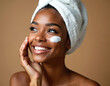 © Vadym - Smiling young african american woman with towel on head applies white cream on cheek. She has clear smooth skin and looks happy enjoying facial treatment.