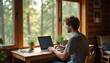 © Vadym - Young man works with laptop in wooden cabin near window overlooking trees. Remote worker types on computer at desk with plants and tablet. Freelancer enjoys tranquil workspace.