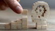© Yuliia - Hand placing wooden block on steps leading to gear-shaped head figure, symbolizing business growth and cognitive development process.