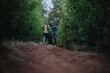 © qunica.com - Two friends hike a dirt forest trail, carrying backpacks under dense green foliage. The scene captures outdoor adventure, fitness, and companionship as they trek through a natural woodland setting.