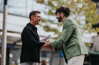 © qunica.com - Two men meet outside in an urban setting, smiling as they shake hands and pass a document. The scene communicates collaboration, agreement, and a casual business moment between friends.