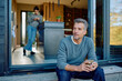 © we.bond.creations - Mature man sitting outside his modern home, holding a cup of coffee and looking away, while a woman uses a phone indoors