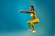 © Prostock-studio - A sporty woman is engaged in deep squats while holding dumbbells, glancing to the side. She is in a blue studio setting that highlights her physical strength and workout routine.