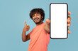 © Prostock-studio - Smiling Indian man with curly hair and beard holds a modern smartphone with a blank display, giving a thumbs up. The background is a bright blue, creating a cheerful atmosphere.