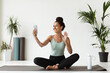 © Prostock-studio - Cheerful young woman sits on a fitness mat in a bright studio, making a video call with her yoga coach. She smiles and waves at her instructor, feeling engaged and motivated during the session.